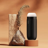 The SVAKOM Sam Neo Vibrating Masturbating Sleeve, a sleek and modern device designed by Svakom, features an elegant, black exterior and sits proudly on a circular base adorned with 5 distinct patterns. Next to it lies a piece of wood and a stone complemented by a decorative dried plant, all set against a warm beige backdrop.